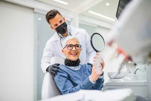 Beautiful senior woman smiling and holding up a mirror to her dental implants at Grins & Giggles Family Dentistry in Spokane Valley, WA