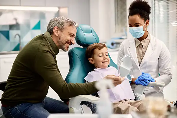 Father holding a mirror for his happy, young son sitting in a dental chair at Grins & Giggles Family Dentistry in Spokane Valley, WA